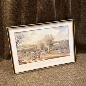 May include: A rectangular framed print titled COURSING AT HATFIELD PARK, featuring a landscape scene with multiple horses, riders, and dogs in an open field. The artwork is enclosed in a thin black and gold frame with a white mat border.