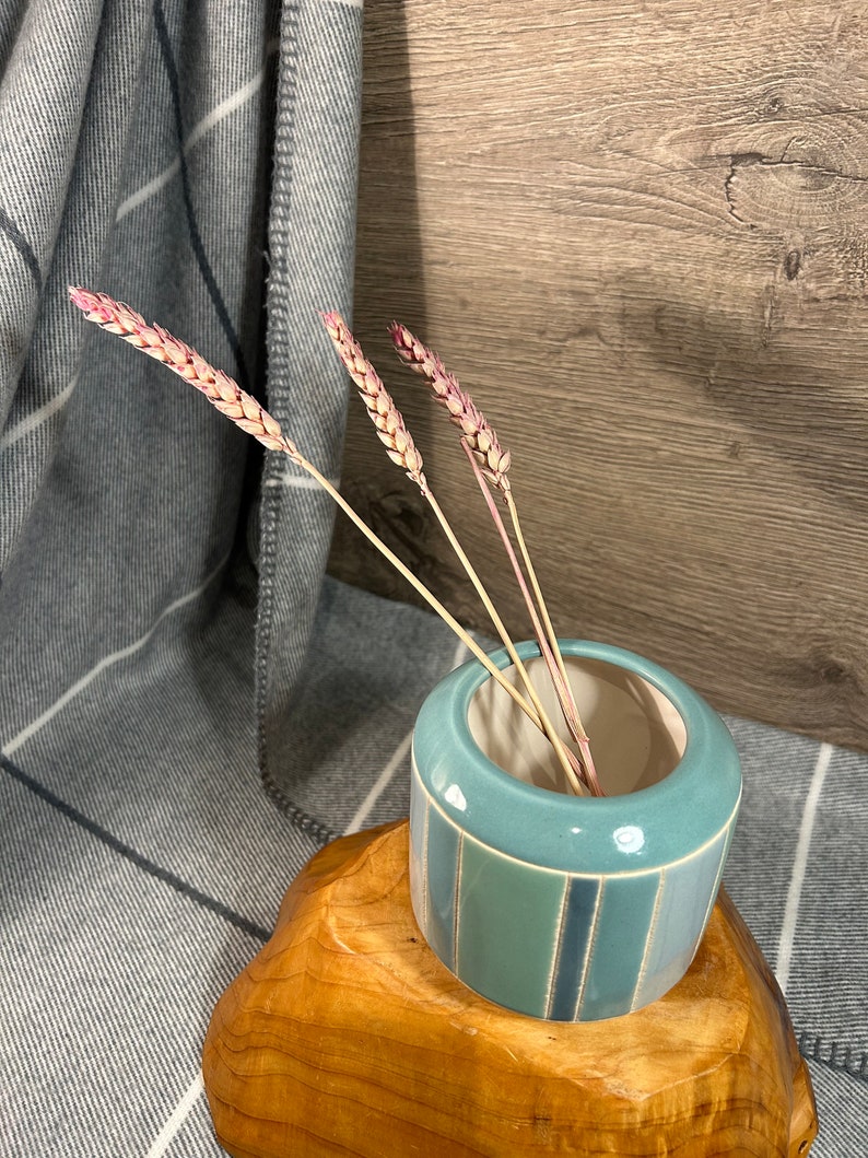 Op de afbeelding: Een lichtblauwe keramische vaas met een gestreept patroon staat op een houten voet. De vaas is gevuld met gedroogde tarwehalmen.