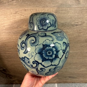 Puede incluir: Un jarrón de cerámica azul y blanco con un diseño floral. El jarrón tiene una tapa y está decorado con un patrón repetitivo de flores y espirales.