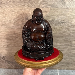 May include: A dark brown Buddha statue with a smiling expression. The statue sits on a dark wooden base with a red velvet cushion, and a gold-colored, round, decorative edge. The statue is a symbol of peace and serenity.