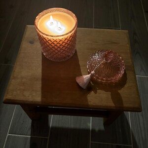 May include: A lit, orange-tinted candle in a cut-glass jar with a lid and tassel sits on a small wooden table. The candle has three wicks and is surrounded by a warm glow. The lid is next to the candle.