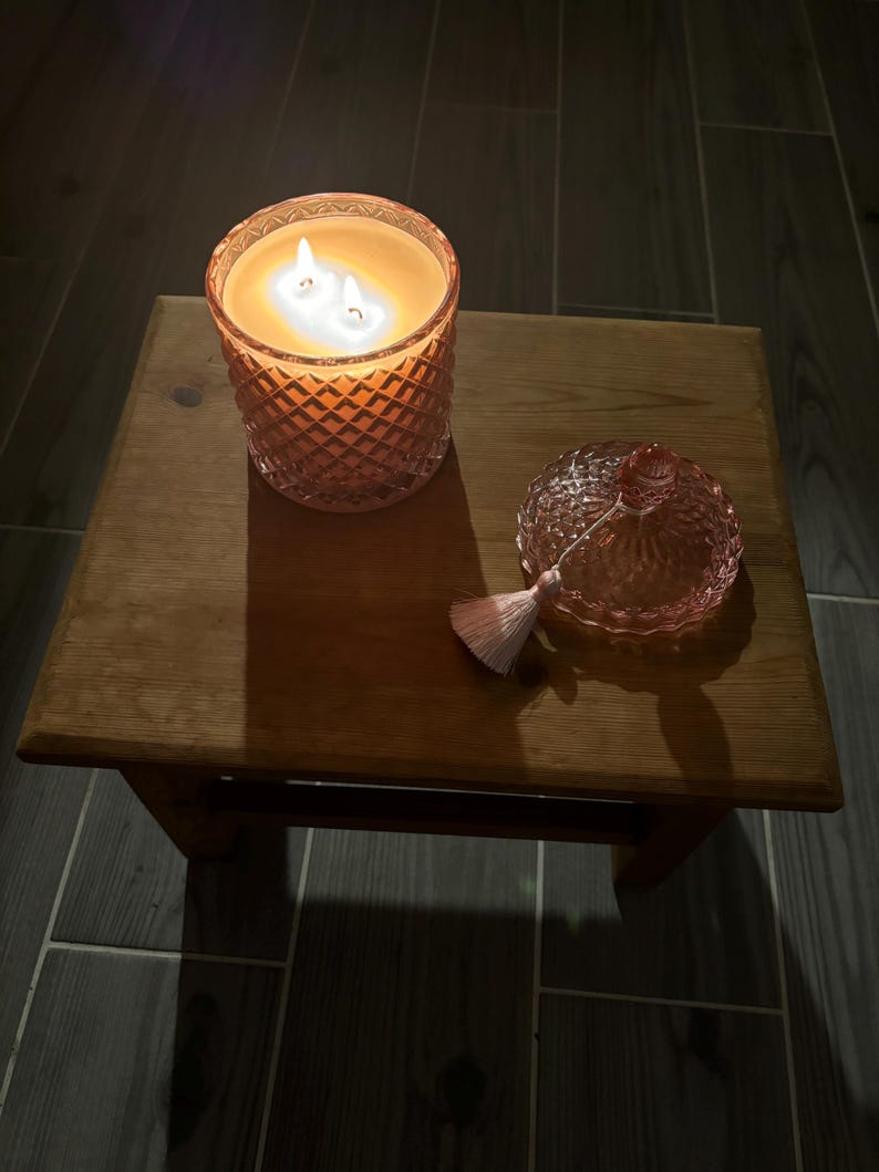 May include: A lit, orange-tinted candle in a cut-glass jar with a lid and tassel sits on a small wooden table. The candle has three wicks and is surrounded by a warm glow. The lid is next to the candle.