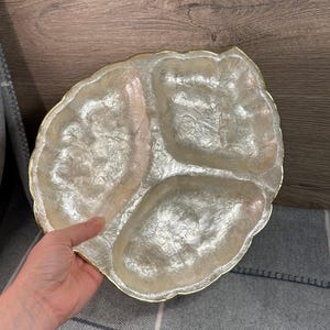 May include: A decorative, three-section serving dish with a pearlescent finish and a gold-colored rim. The dish has a leaf-like shape and is designed for serving appetizers or snacks. The dish is held by a hand.