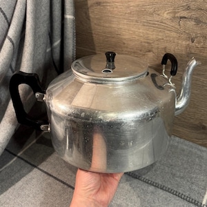 May include: A vintage aluminum teapot with a lid and a curved spout. The teapot has a black handle and a black knob on the lid. The teapot is sitting on a gray and white plaid blanket.