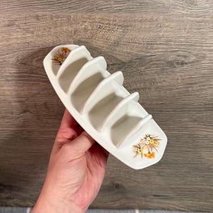 Puede incluir: Un tostador de cerámica blanca con cuatro ranuras. El soporte tiene forma curva y está decorado con diseños florales en amarillo, naranja y marrón. El soporte está sostenido por una mano.