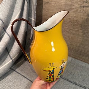 Puede incluir: Una jarra de esmalte amarillo con un asa marrón y un interior blanco. La jarra presenta dos ilustraciones de jirafas con manchas coloridas. La jarra es sostenida por una persona.