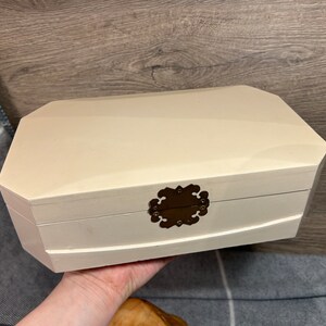 May include: A white wooden jewelry box with a decorative brass clasp. The box has a slightly rounded shape and a smooth finish.