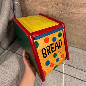 May include: A hand-painted wooden bread bin with a yellow lid, green sides, and a front panel featuring the word "BREAD" and colourful polka dots. The box has red and blue trim.