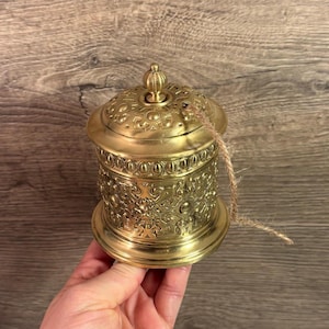 Puede incluir: Un recipiente dorado y ornamentado, con tapa abovedada y complejos diseños florales. El recipiente tiene un cuerpo cilíndrico y un pequeño pomo decorativo en la parte superior. Una cuerda de fibra natural está atada al costado.