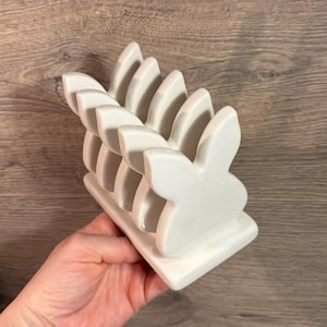 Puede incluir: Un servilletero de cerámica blanca con forma de conejito. El soporte tiene una base rectangular y una serie de siluetas de conejos para sujetar las servilletas. El diseño es sencillo y elegante, adecuado para la decoración de Pascua o primavera.