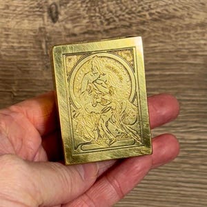 May include: A gold-colored metal box with an engraved design of a woman in a flowing gown. The box has a slightly textured surface.
