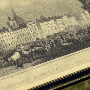 May include: An antique black and white print of Smithfield Market, From the Bars. The print depicts a bustling market scene with buildings, people, and livestock. The print is framed in a dark blue frame with a gold trim.