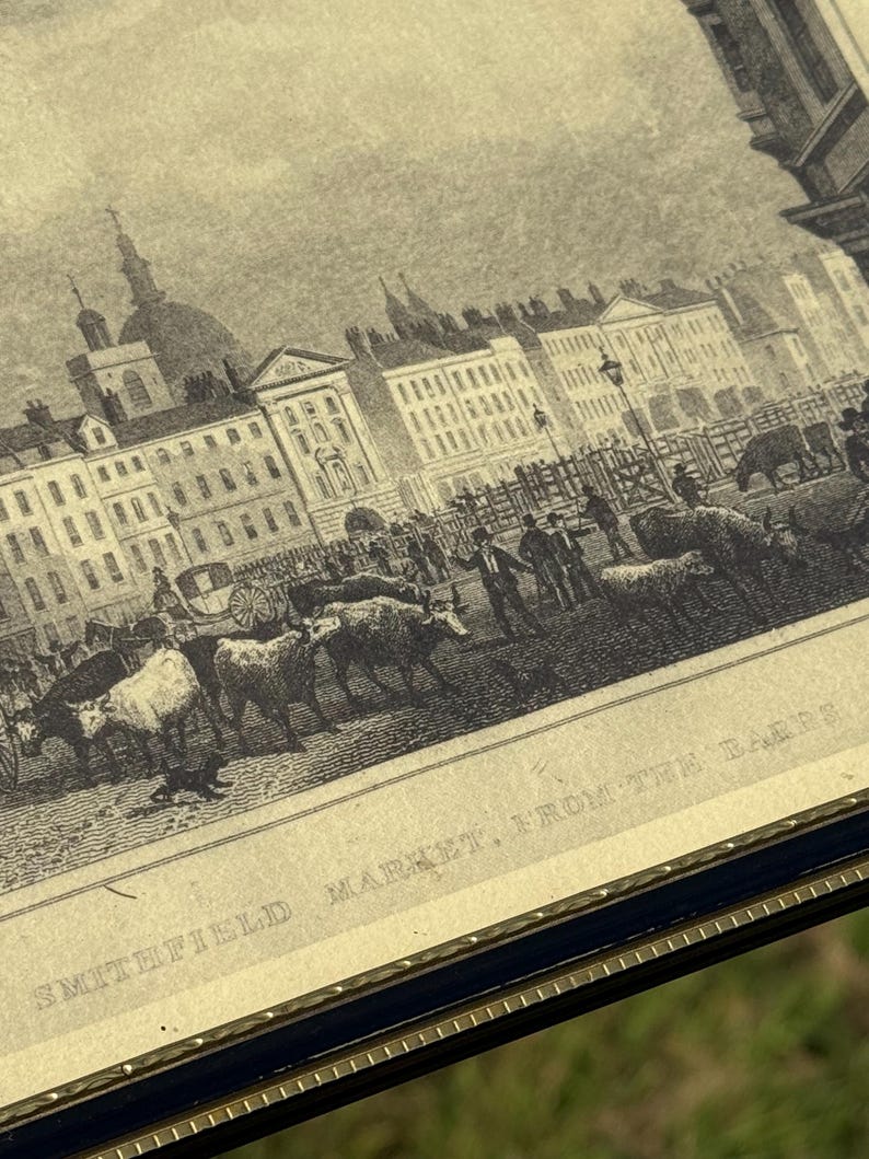 May include: An antique black and white print of Smithfield Market, From the Bars. The print depicts a bustling market scene with buildings, people, and livestock. The print is framed in a dark blue frame with a gold trim.