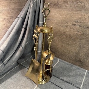 Puede incluir: Un juego de herramientas para chimenea de latón con soporte. El juego incluye una pala, un atizador y un cepillo. Los mangos tienen recortes en forma de corazón. Las herramientas están dispuestas sobre un soporte con base circular. El fondo presenta una manta a cuadros gris y blanca y una superficie de madera.