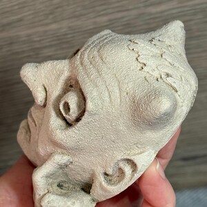 May include: A small, off-white sculpture of a face with detailed features, including a prominent nose, curled ear, and textured surface. The sculpture has a slightly weathered appearance.