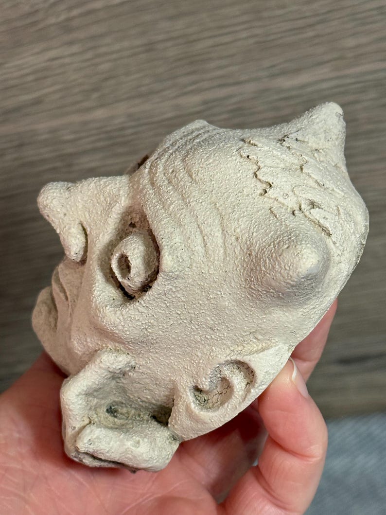 May include: A small, off-white sculpture of a face with detailed features, including a prominent nose, curled ear, and textured surface. The sculpture has a slightly weathered appearance.