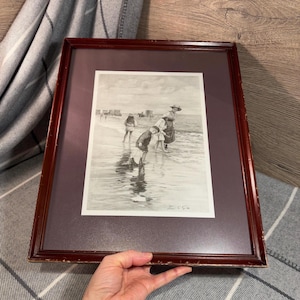 May include: A framed black and white print depicting a family at the beach. The print is set in a dark brown wooden frame with a purple mat. The artwork shows a mother and children playing in the water.