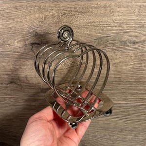 Puede incluir: Un tostador de metal plateado en forma de corazón. El soporte tiene una base con tres pequeños pies de bola y un remolino decorativo en la parte superior. Diseñado para contener varias rebanadas de tostadas.
