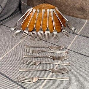 Può includere: Una collezione di forchette d'argento disposte su una superficie a scacchi grigia e bianca. Alcune forchette sono poste attorno a una ciotola di legno, mentre altre sono distese. Le forchette hanno un design classico.