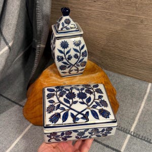 May include: Two ceramic decorative pieces with a white base and blue floral design. One is a small lidded jar, and the other is a rectangular box with a lid. Both have gold trim. The pieces are on a wooden surface.