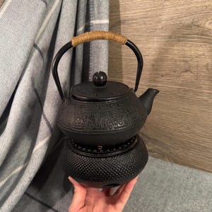 Könnte beinhalten: Schwarze Gusseisen-Teekanne mit dekorativem Muster und geflochtenem Griff. Die Teekanne steht auf einem passenden Wärmer. Der Griff ist mit einem beigefarbenen Material umwickelt. Die Teekanne ist etwa 15 cm hoch.