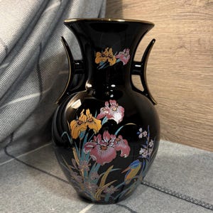 Op de afbeelding: Zwarte keramische vaas met gouden accenten en een bloemenontwerp met gele, roze en blauwe bloemen. Een kleine blauwe vogel zit op een tak.