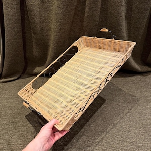 May include: A rectangular serving tray featuring a woven light brown wicker base and sides. The tray is framed by a black metal structure with decorative scrollwork along the sides and two small black metal handles on the ends.