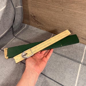 Könnte beinhalten: Ein Vintage-Rechenschieber mit beigem Gehäuse und schwarzen Markierungen, teilweise aus einem grünen Etui herausgezogen. Der Rechenschieber hat ein kreisförmiges Zifferblatt. Er wird vor einer grau-weiß karierten Decke gehalten.