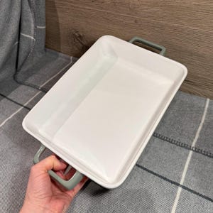May include: A white rectangular ceramic baking dish with rounded corners and two grey handles. The dish is empty and rests on a grey and white plaid blanket. The background is a wooden surface.
