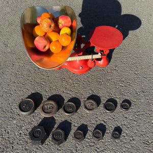 May include: A vintage red Avery scale with a silver bowl filled with oranges, apples, and a lemon. Weights of various sizes are arranged below the scale, including 2lb, 1lb, and 4oz weights. The scale is on a gray surface.