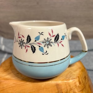 Puede incluir: Una jarra de crema blanca con un borde azul y dorado y un diseño floral en negro, rosa y azul.