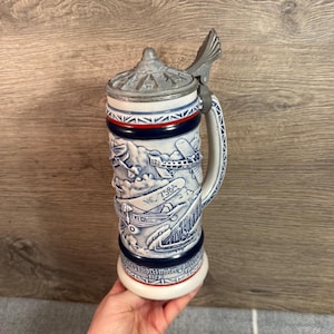 May include: A ceramic beer stein with a hinged metal lid. The stein is white with blue and red accents and features a detailed relief design of boats, planes, and the text "NC 7584". The base reads "Tar Heel Motor Boat 1977".