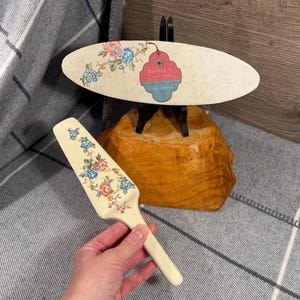 May include: A cream-coloured cake server with a floral design in pink and blue. The server is held in a hand. A matching cake stand with the text "Rosie's Cakes" is in the background.