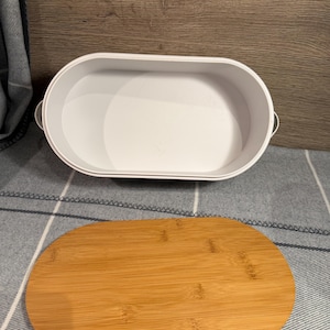May include: An oval-shaped white container with small side handles and a separate, matching oval-shaped bamboo lid. The container has a smooth, solid white interior and exterior, while the lid features a natural light brown wood grain finish.