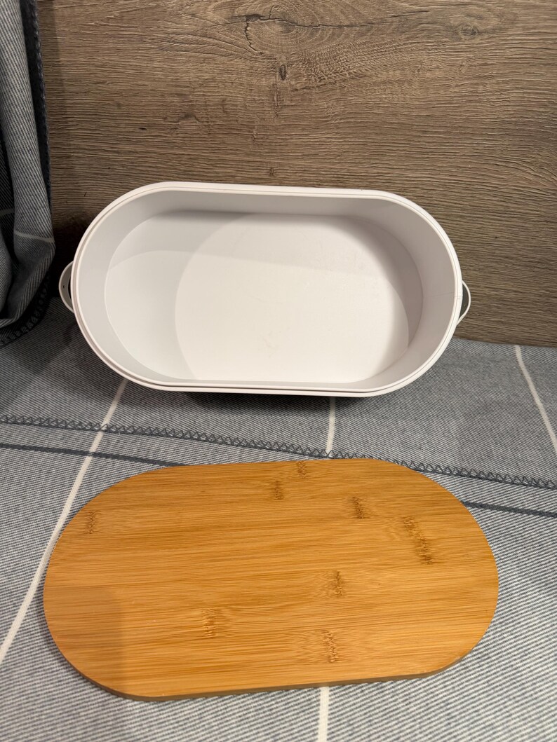 May include: An oval-shaped white container with small side handles and a separate, matching oval-shaped bamboo lid. The container has a smooth, solid white interior and exterior, while the lid features a natural light brown wood grain finish.