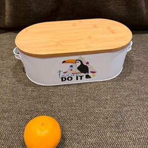 May include: An oval white metal storage container with a light brown bamboo lid and two side handles. The front features a graphic of a toucan with colorful stars and the text "toucan DO IT". A single round orange fruit is placed in front of the container.