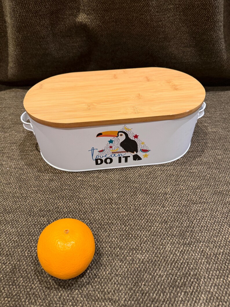 May include: An oval white metal storage container with a light brown bamboo lid and two side handles. The front features a graphic of a toucan with colorful stars and the text "toucan DO IT". A single round orange fruit is placed in front of the container.
