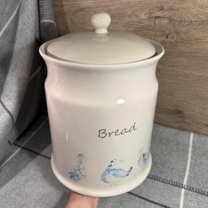 May include: A white ceramic bread bin with a lid and a small knob. The word "Bread" is written on the front in a script font. The bottom of the bin features blue duck illustrations.
