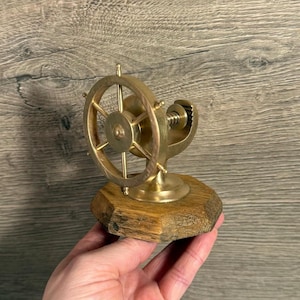 May include: A brass ship's wheel nutcracker mounted on a wooden base. The nutcracker features a detailed wheel design with spokes and a central hub. The wooden base has a natural, rustic appearance.
