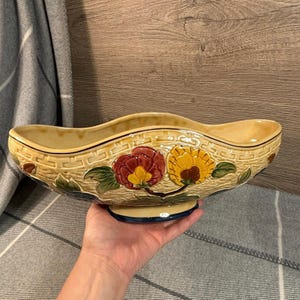 Puede incluir: Un cuenco decorativo de cerámica con un diseño texturizado de tejido de cesta. El cuenco es de color beige con un borde ondulado y una base de pedestal. Presenta diseños florales en rojo, amarillo y verde. El cuenco mide unos 30 cm de largo.