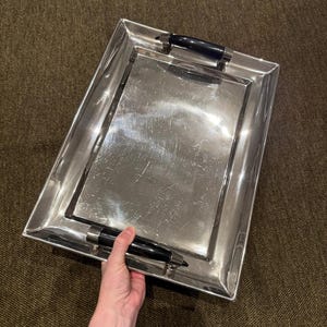 May include: A rectangular silver serving tray with black handles. The tray has a polished, reflective surface and a raised border. The black handles are cylindrical and attached to the tray's frame. The tray is held by a person's hand.