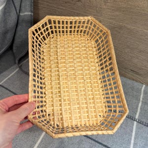 May include: A rectangular woven basket in light brown. The basket has a geometric shape with a woven pattern. The basket is held by a hand and is placed on a grey and white plaid blanket.
