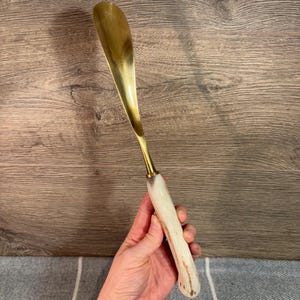 May include: A gold shoehorn with a white and brown antler handle.