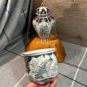Könnte beinhalten: Ein weißer Keramik-Pflanztopf mit goldenem Rand und blau-goldener Zierbordüre. Der Pflanztopf hat ein farbenfrohes Pfauen-Design. Ein passendes Gefäß mit Deckel und Pfauen-Design steht hinter dem Pflanztopf.