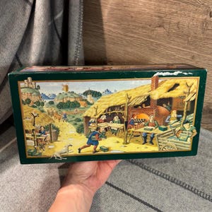 May include: A rectangular, dark green box with a detailed, colorful illustration of a medieval village scene. The artwork depicts people working, a dog, and buildings with thatched roofs. The box is held by a person.