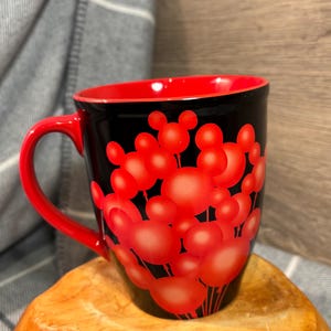 Peut inclure: Mug &agrave; caf&eacute; noir avec int&eacute;rieur et anse rouges. Le mug est orn&eacute; de formes de t&ecirc;tes de Mickey Mouse rouges sur fond noir. Le mug est pos&eacute; sur une surface en bois.