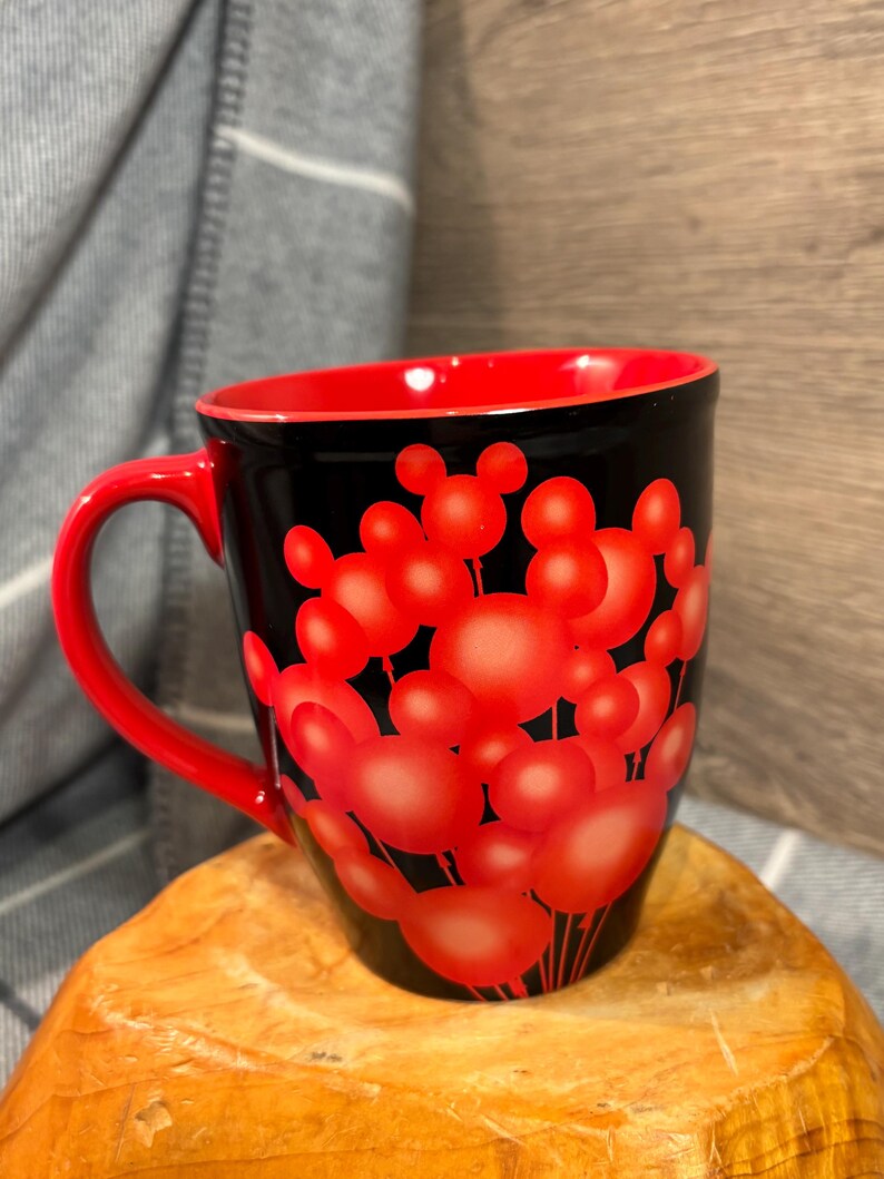 Peut inclure: Mug &agrave; caf&eacute; noir avec int&eacute;rieur et anse rouges. Le mug est orn&eacute; de formes de t&ecirc;tes de Mickey Mouse rouges sur fond noir. Le mug est pos&eacute; sur une surface en bois.