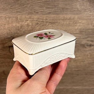 Op de afbeelding: Een witte porseleinen sieradenkistje met een bloemenmotief op de deksel. De kist heeft een gouden rand en decoratieve pootjes.