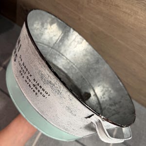 Op de afbeelding: Een ronde metalen kom met een lichtgroene basis en een verweerde witte bovenrand. Het witte gedeelte is voorzien van zwarte tekst: De beaux oiseaux volent au-dessus des arbres. De kom is uitgerust met twee kleine metalen handgrepen aan de zijkant.