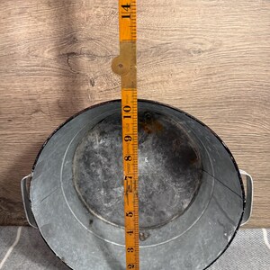 Op de afbeelding: Een ronde gegalvaniseerde metalen kom met twee handgrepen aan de zijkant en een donkere rand. Een gele opvouwbare houten liniaal ligt in het midden en toont een diameter van ongeveer 26,5 cm. De binnenkant vertoont sporen van slijtage.
